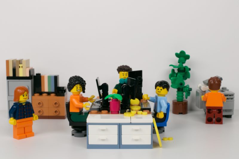 A Lego minifigure office scene: colleagues at desks with a computer, filing cabinet, bookshelf and pot plant.